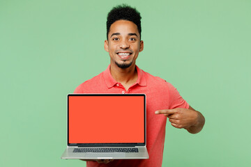 Wall Mural - Young IT man of African American ethnicity wears pink t-shirt hold use work point finger on laptop pc computer with blank screen workspace area isolated on plain green background. Lifestyle concept.