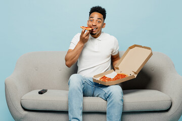 Wall Mural - Full body young man fan wear t-shirt cheer up support football sport team hold soccer ball italian pizza in flatbox look camera sit on grey sofa watch tv live stream isolated on plain blue background.