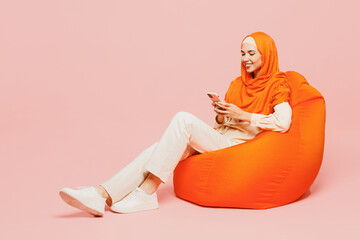 Wall Mural - Full body young arabian asian muslim woman wears orange abaya hijab sit in bag chair hold in hand use mobile cell phone isolated on plain pink background. Uae middle eastern islam religious concept.