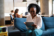 © JustLife - Beautiful African businesswoman sitting on sofa in office. Young woman using digital tablet.