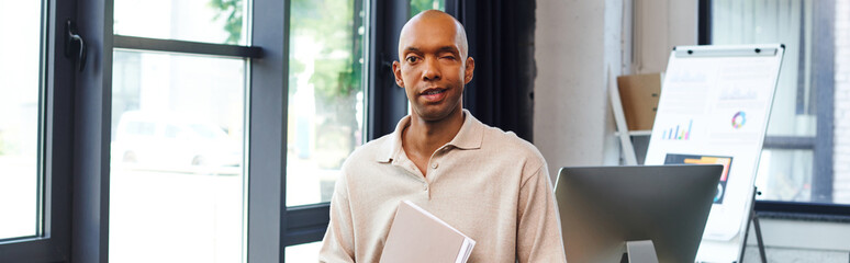 Wall Mural - dark skinned man with myasthenia gravis disease holding folder and standing in office, bold african american worker looking at camera, inclusion, monitor and graphics, banner