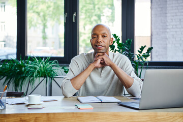 Wall Mural - ptosis syndrome, bold african american businessman with eye syndrome looking at camera near laptop on desk, myasthenia gravis, cheerful dark skinned office worker, diversity and inclusion