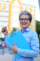 Sticker - Smiling woman realtor with keys and documents in her hands against the backdrop of a multi-storey building.