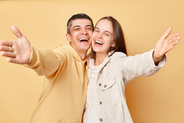 Wall Mural - Friendly smiling woman and man wearing casual style clothing standing isolated over beige background spreading hands inviting showing welcome gesture laughing with happiness.