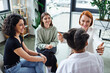 © LIGHTFIELD STUDIOS - happy redhead motivation coach sitting with outstretched hands and talking to diverse group of multiethnic women during therapy in consulting room, friendship and mental wellness concept