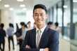 © SayLi - portrait of a handsome smiling asian businessman boss in a suit standing in his modern business company office. his workers standing in the blurry background. Generative AI