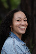 © Levi Meir Clancy - A multiracial woman with long, curly black hair and a joyful, beaming smile turns to make eye contact with the camera. She wears big hoop earrings and a denim jacket. She is happy and confident.