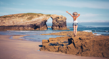 Wall Mural - Woman standin on rock enjoying natural arch in atlantic ocean- Galicia in Spain- Cathedrals beach