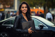 © J S - Beautiful Indian female real estate agent or businesswoman smiling and holding a leather document folder with copy space on the blurred off focus background of cars and traffic