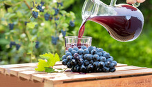 Fresh Grape Juice Pouring Into Glass From Jug With Ripe Bunch Of Dark Blue Berries On Crate As Table In The Garden