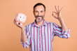 Portrait of cheerful handsome person toothy smile arm hold money pig bank demonstrate okey symbol isolated on beige color background