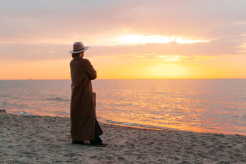 Lonely pensive woman in a coat and hat standing at sunset near sea, enjoying nature. Back view, copy space