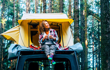 Camping and vehicle car with roof tent outdoors leisure activity and tourism lifestyle people. One woman sitting outside her tent and admire forest green outside having relax and chilling. Vacation