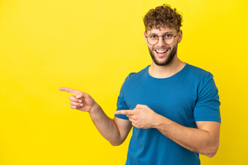 Poster - Young handsome caucasian man isolated on yellow background surprised and pointing side