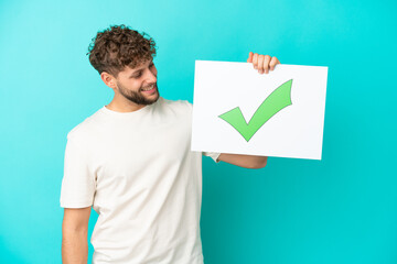 Wall Mural - Young handsome caucasian man isolated on blue background holding a placard with text Green check mark icon with happy expression