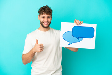 Wall Mural - Young handsome caucasian man isolated on blue background holding a placard with speech bubble icon with thumb up