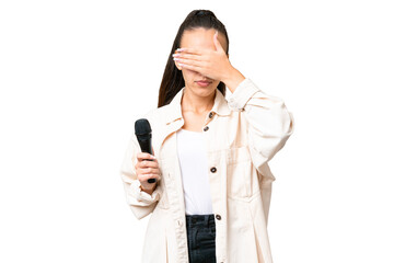 Wall Mural - Young singer woman picking up a microphone over isolated chroma key background covering eyes by hands. Do not want to see something