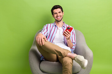 Poster - Full size photo of handsome positive person wear stylish clothes sit in armchair hold smartphone isolated on green color background