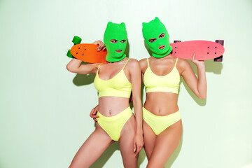 Two beautiful sexy women in green underwear. Models wearing bandit balaclava mask. Hot seductive female in nice lingerie posing near blue wall in studio. Crime and violence. Hold penny skateboard