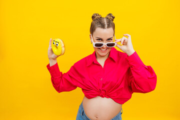 Cheerful pregnant woman holding a sweet yellow donut in her hand on a yellow background. Expecting a child, pregnancy and motherhood. The concept of healthy and unhealthy food, diet. Junk food