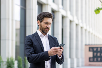 Serious thinking businessman boss holding phone, man reading online message and typing, investor outside office building, successful financier concentrated banker, using app on smartphone