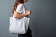 A woman is carrying a canvas tote bag. The tote bag mockup is white with grey background
