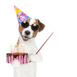 Jack russell terrier puppy  wearing sunglasses and party cap holds birthday cake with burning candles and points away on empty space. Isolated on white background