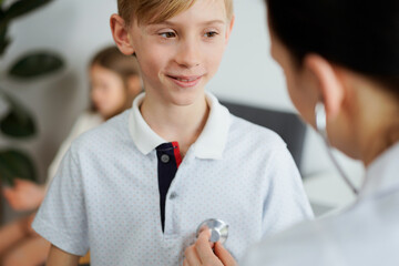 Wall Mural - Doctor and kid patient are in the clinic. Physician in white coat examining a smiling young boy with a stethoscope, close up. Medicine, therapy concept