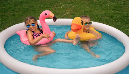 Two funny little girls in sunglasses are swimming in circles in a small inflatable swimming pool in the yard. Water fun on vacation