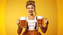 Young Sexy Oktoberfest Waitress, Wearing A Traditional Bavarian Dress, Serving Big Beer Mugs On Yellow Background
