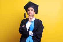 Thoughtful Face Of Young Male Wearing Graduation Cap And Ceremony Robe Over Yellow Background With Hand On Chin, Looking Up, Thinking About Question, Pensive Expression, Looking For An Idea