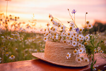 A Straw Hat And A Bouquet Of Wild Flowers On A Wooden Bench In A Field At Sunset On A Summer Evening.