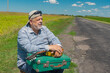 Portrait of bearded Ukrainian senior in black sunglasses sitting  on roadside  with an ancient green suitcase and mandolin at summer season