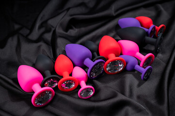 A set of silicone anal plugs in different colors and sizes on a black silk sheet. 