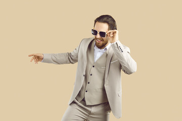 Happy man in formal suit having fun in fashion studio. Cheerful joyful carefree young businessman wearing custom tailored three piece suit and cool sunglasses dancing isolated on beigebackground