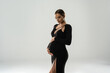 Pregnancy concept. Woman posing in studio wearing black elegant dress, holding her pregnant belly, waiting for a baby.