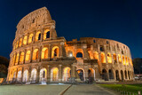 Fototapeta Londyn - Nighttime photo of the Roman Colosseum. The structure is lit up with a warm orange glow set agains a cobalt blue sky. No people are visible.
