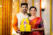 © WESTOCK - Happy young indian couple with gift boxes for diwali festival celebration looking camera at home - concept of festival sale, offers and surprise presents for shopping.