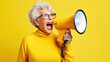 © MP Studio - An elderly woman screams into a loudspeaker against a yellow background.