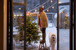 Young woman decorates Christmas tree, attaching festive bow on top while standing on step ladder at beautiful snowy backyard with dog, view through the window. Preparation for the winter holidays