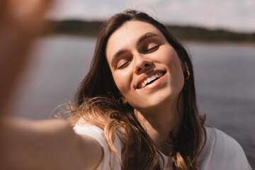 Wall Mural - Excited brunette woman in white t shirt smiles with closed eyes. Girl takes selfie walking on the lake background. Happy smiling emotions, relaxation.