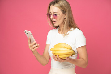 Attractive woman is ordering products online concept. beautiful woman in eyewear with concentraited face reading information on smartphone and holds yellow bananas in hand isolated on pink background