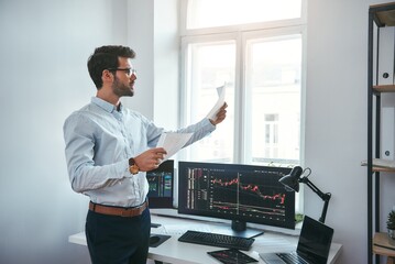 Fototapete - Trading strategy. Smart and young trader in eyeglasses looking at financial reports and analyzing trading charts while standing in front of computer screens in modern office.