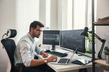 Fototapete - Busy working day. Young bearded trader in eyeglasses working with laptop while sitting in his modern office in front of computer screens with trading charts.