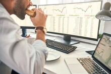Enjoying Lunch. Close-up Of Bearded Trader Is Eating Fresh Sandwich And Looking At Monitor Screen With Financial Data While Sitting In His Modern Office