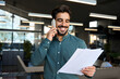 © insta_photos - Smiling happy young bearded Latin professional business man executive holding documents and cell phone making mobile call at work on cellphone consulting client standing in modern office.