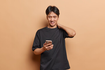 Wall Mural - People positive emotions. Young cheerful smiling Asian man wearing casual black tshirt standing in centre on beige background holding smartphone reading funny messages or information on internet