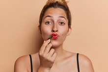 Beauty and cosmetic. Studio shot of young pretty glad European lady applying red lipstick standing in centre isolated on beige background looking straight at camera keeping long brunette hair in bun