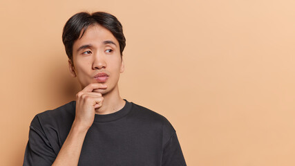 Wall Mural - People emotions concept. Young thoughtful Asian man holding left hand on chin trying to solve difficult task or problem standing on left isolated on beige background with blank space for advertisement