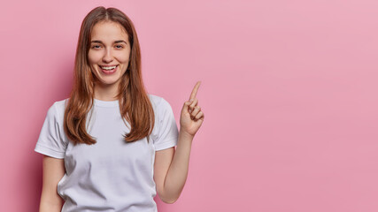 Wall Mural - Horizontal shot of pleased cheerful woman with straight hair points index finger on blank space dressed in casual white t shirt shows advetisement isolated ove pink background. Look at this.